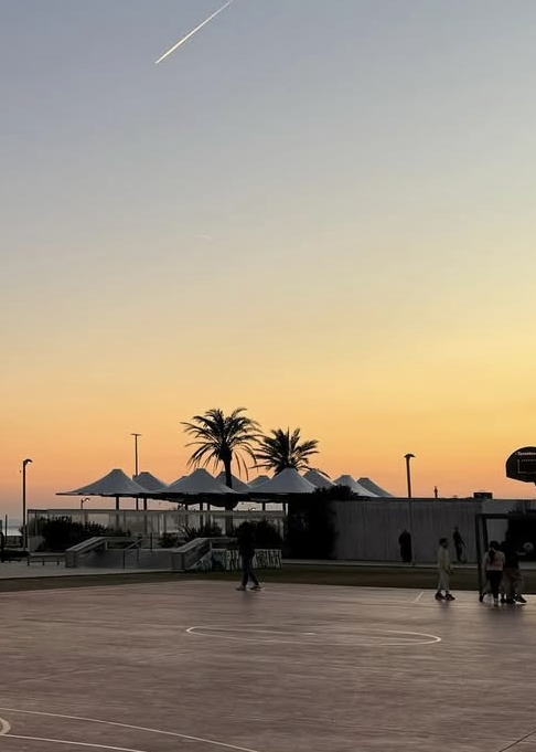 Sonnenuntergang auf einem Basketballplatz mit spielenden Leuten, Palmen im Hintergrund und einem Flugzeug. 